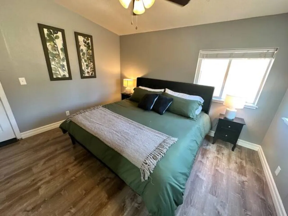 This serene interior scene showcases a cozy bed and nightstand next to expansive windows, with natural light flooding the room. The view outside the windows hints at lush greenery, and the overall arrangement creates an ideal nook for reading or relaxing while feeling connected to the outdoors.