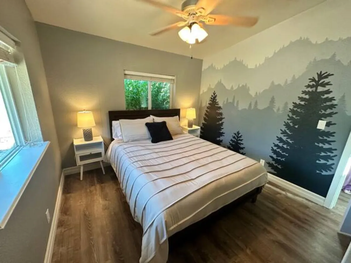 This serene bedroom features a cozy bed set against a striking mountain mural, complemented by a nightstand with a warm lamp and a window letting in natural light. The overall mood is tranquil, inviting rest and relaxation.