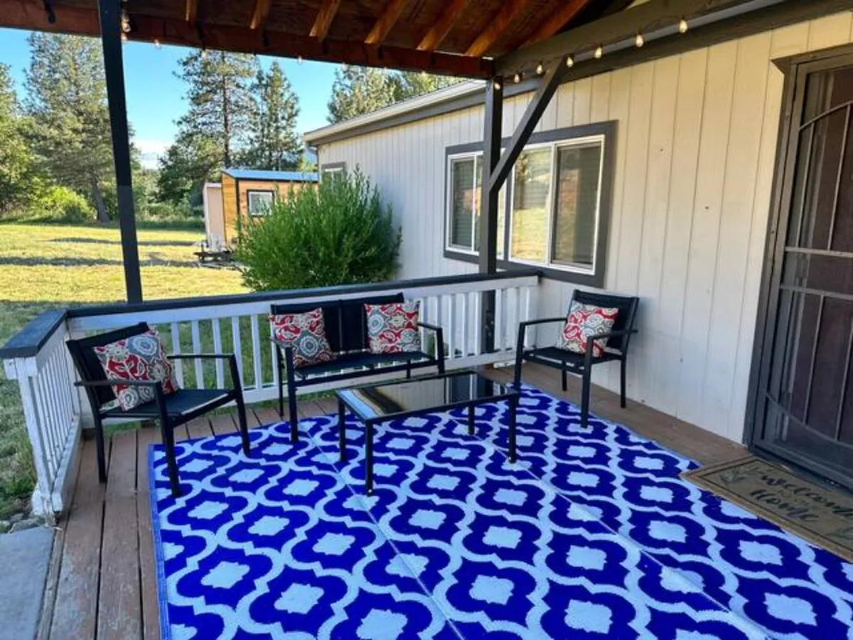 An inviting exterior porch scene features a colorful outdoor rug, cozy seating, and is surrounded by lush greenery. A book and a mug suggest a spot ideal for reading and relaxation, with the open-air setting blurring the line between inside comfort and outside beauty.