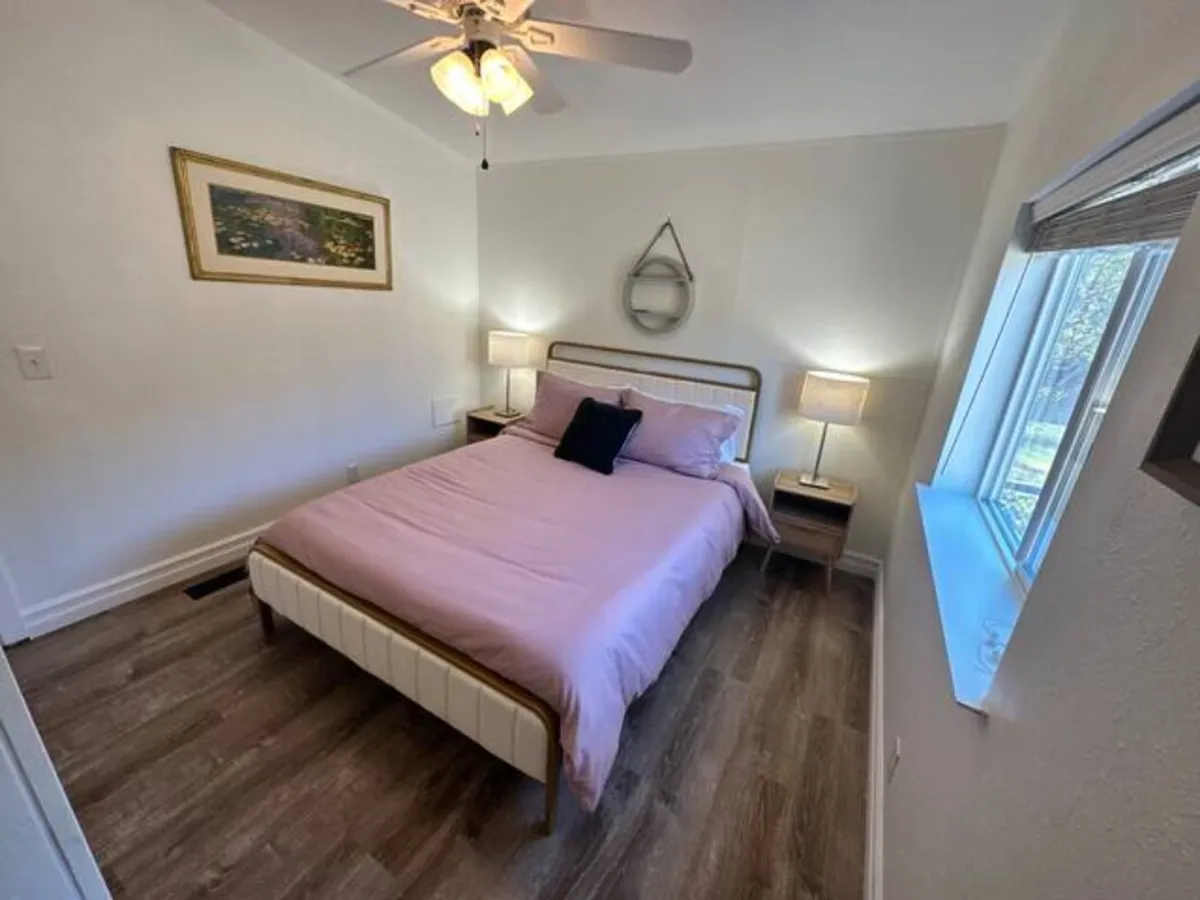 A welcoming bedroom with soft pink bedding, twin bedside lamps, framed wall art, and a window streaming in gentle morning sunlight. The space encourages relaxation, reading, and cherished quiet moments.