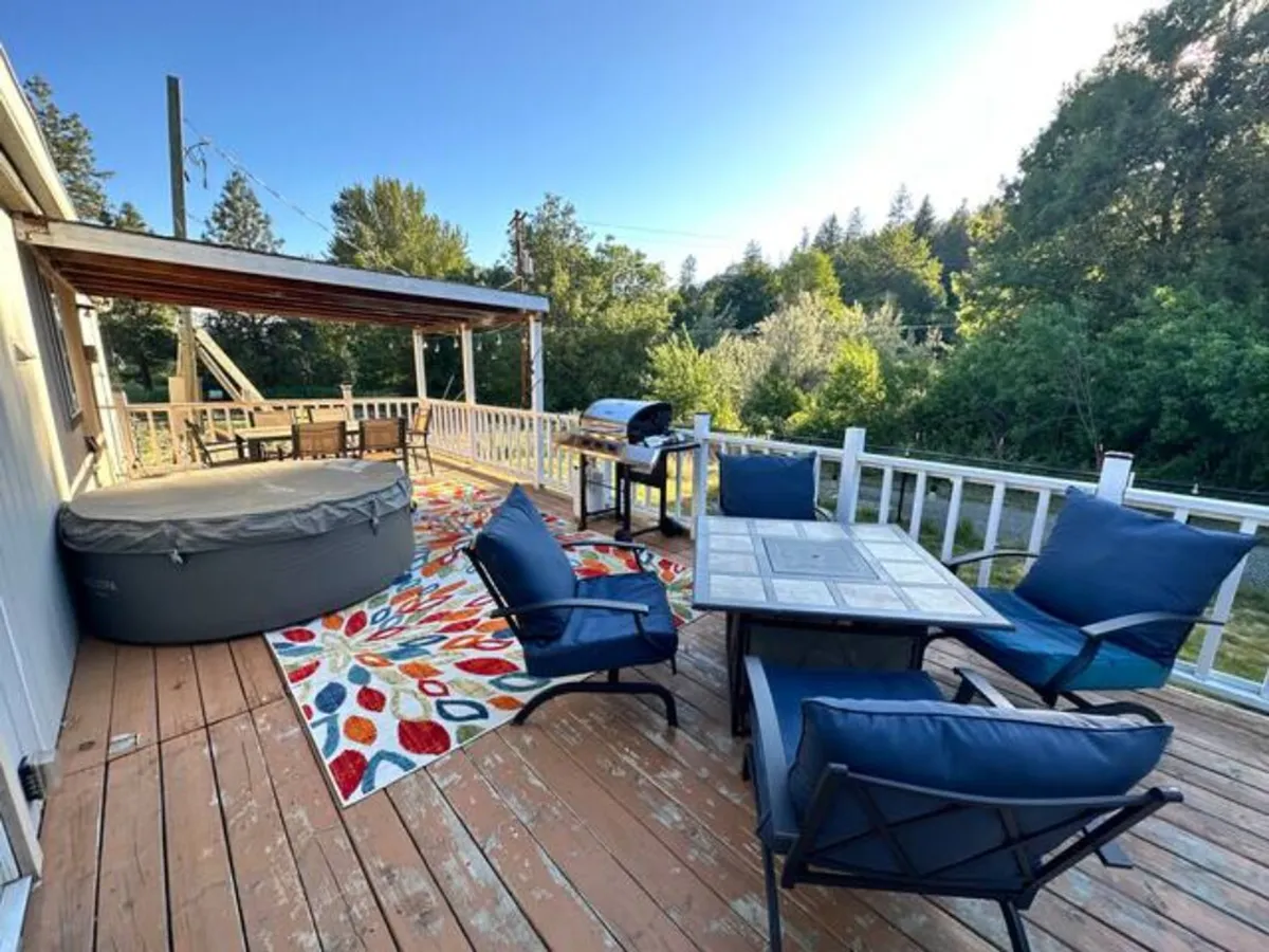 This image features an inviting farmhouse exterior space with a hot tub, outdoor seating area, a grill, and a colorful rug. The setting is surrounded by trees and open sky, blending the comfort of the retreat with the natural landscape. The outdoor area is set up for relaxation and social activities, with a direct connection to the surrounding nature.