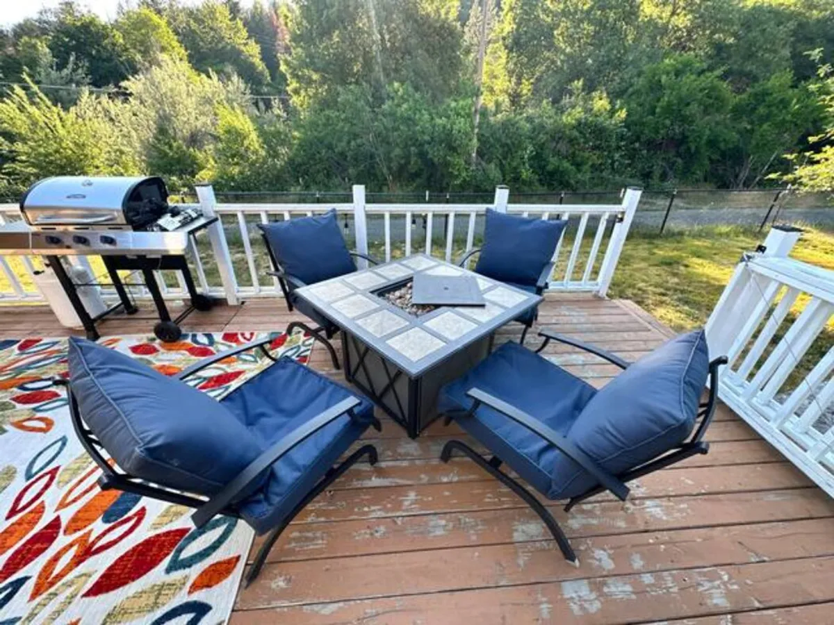 This exterior shot showcases a welcoming deck area with a fire pit table, comfortable seating, a grill, and vibrant outdoor rug. The setup invites guests to relax outdoors, with views of the surrounding landscape that hint at the proximity to nature trails and peaceful woodlands.