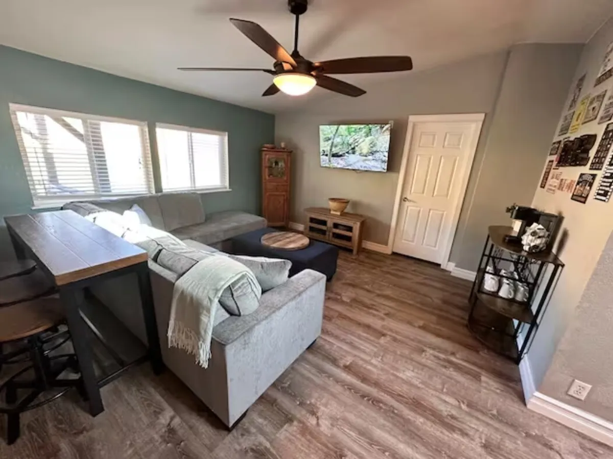 This image presents a cozy living area with a sofa, coffee table, television, windows, and a bar area. The spatial arrangement and proximity to a bar area suggest this space is adjacent to or visible from the entryway, giving guests an immediate sense of warmth and relaxation upon entry. Large windows allow for natural light, enhancing the inviting mood.