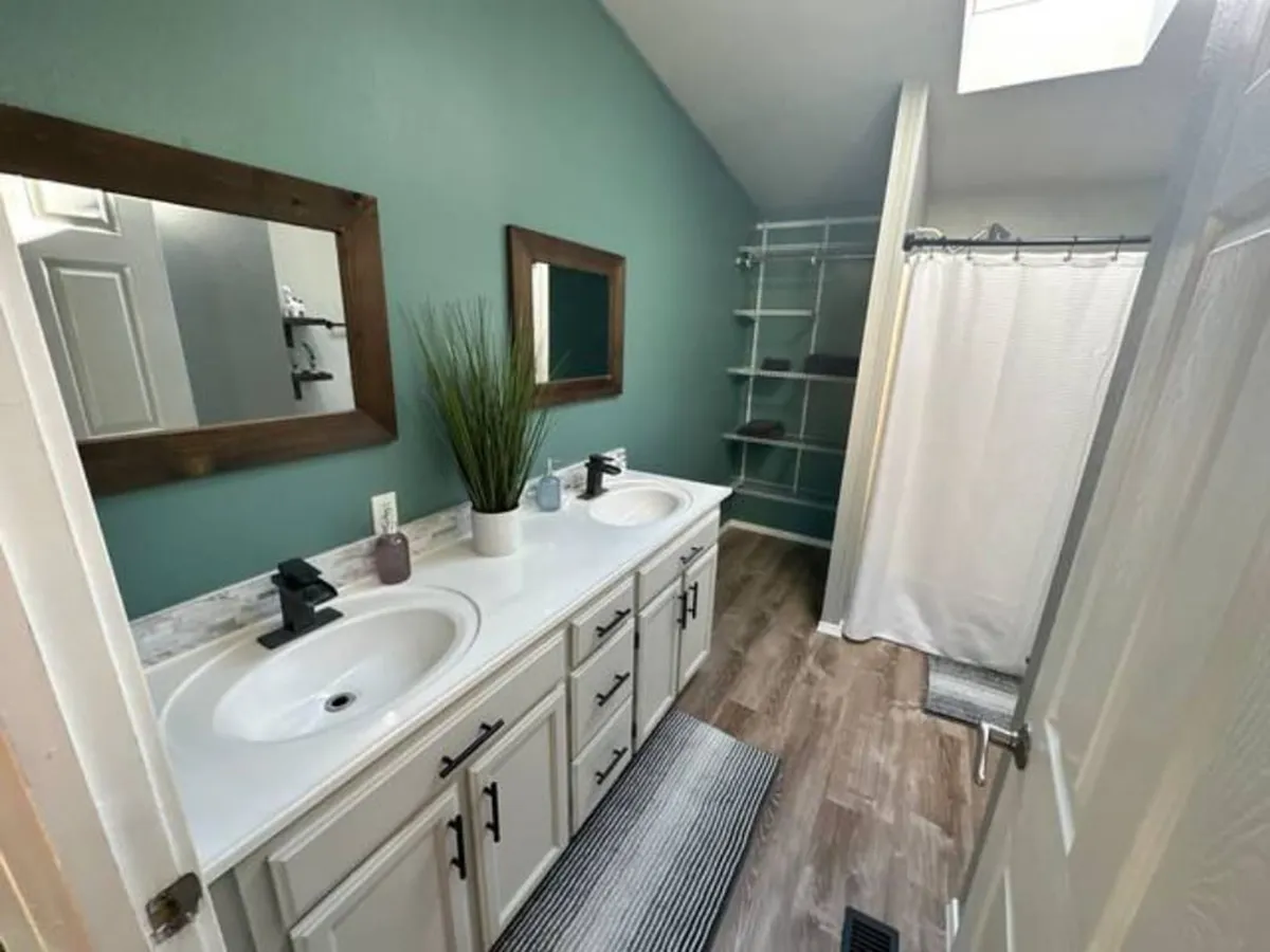 A stylish bathroom featuring a double sink vanity, elegant mirrors, a spacious shower area, open shelving, and a decorative plant. The space radiates calm and freshness, perfect for morning or evening routines.